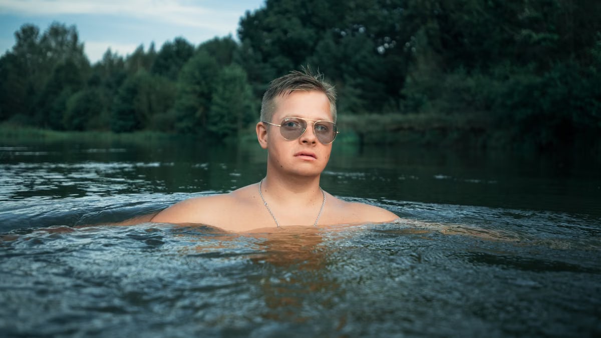 Man with sunglasses enjoys a refreshing swim in a river surrounded by lush greenery.